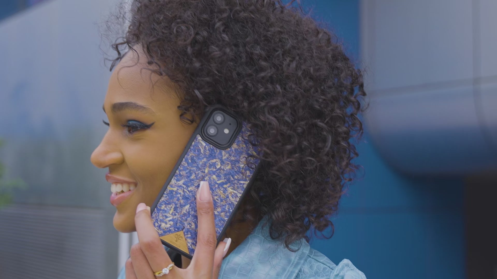 Une très jolie femme souriante et capverdienne au regard charismatique tiens son téléphone contre son oreille. Son téléphone est vêtu d’une coque Hibisca. La coque est exceptionnelle. Elle a pour façade des fleurs de bleuets compressées très jolie à voir. La coque est donc de couleur bleu.. La femme tenant le téléphone a une belle main avec un vernis artistique sur ses ongles. Elle porte également un manteau bleu élégant avec un effet peau de crocodile avec une tenue moulante noire à l’intérieur.