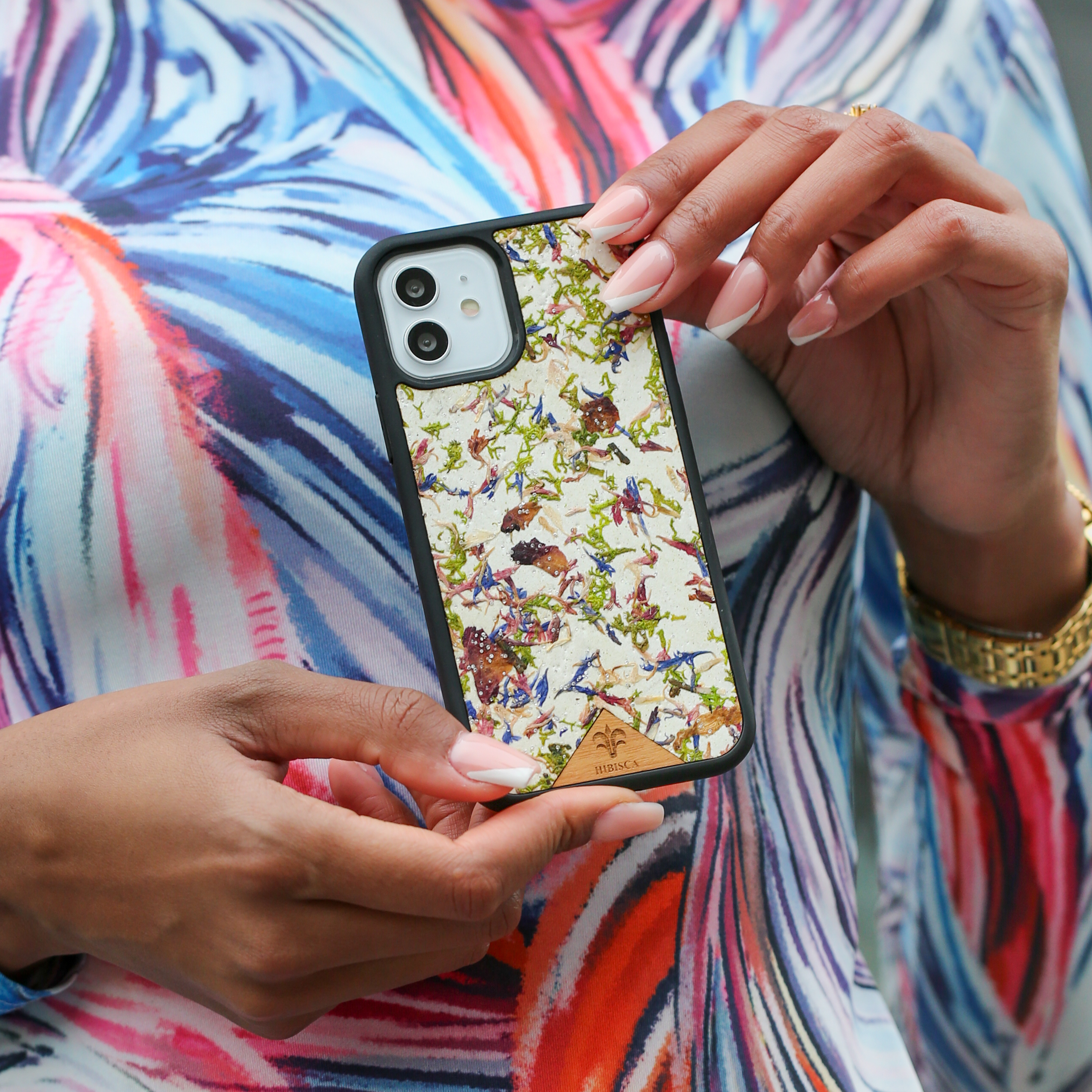 Une jolie femme capverdienne tient son téléphone d’une coque fleurie Hibisca. Elle a pour façade de multiples fleurs. Nous pouvons y apercevoir du blanc et de multiples cristaux de chez Swarovski. La femme a de belles mains avec un vernis artistique sur ses ongles et porte une belle robe moulante avec des jeux de teintes rouge, blanc, orange, bleu et violet.