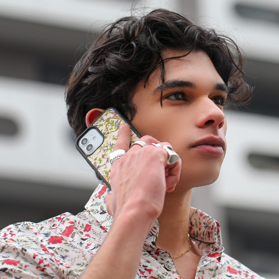 Un très bel homme au regard fier tient son téléphone contre son oreille. Son téléphone est vêtu d’une coque fleurie Hibisca. La coque fleurie est exceptionnelle. Elle a pour façade de multiples fleurs. Nous pouvons y apercevoir du blanc. Elle est aussi parsemée de multiple cristaux de chez Swarovski. L’homme tenant le téléphone a une belle main ornée de bague sur chacun de ses doigts. Il porte également une très jolie chemise blanche avec des motifs fleuris rouge et verts.