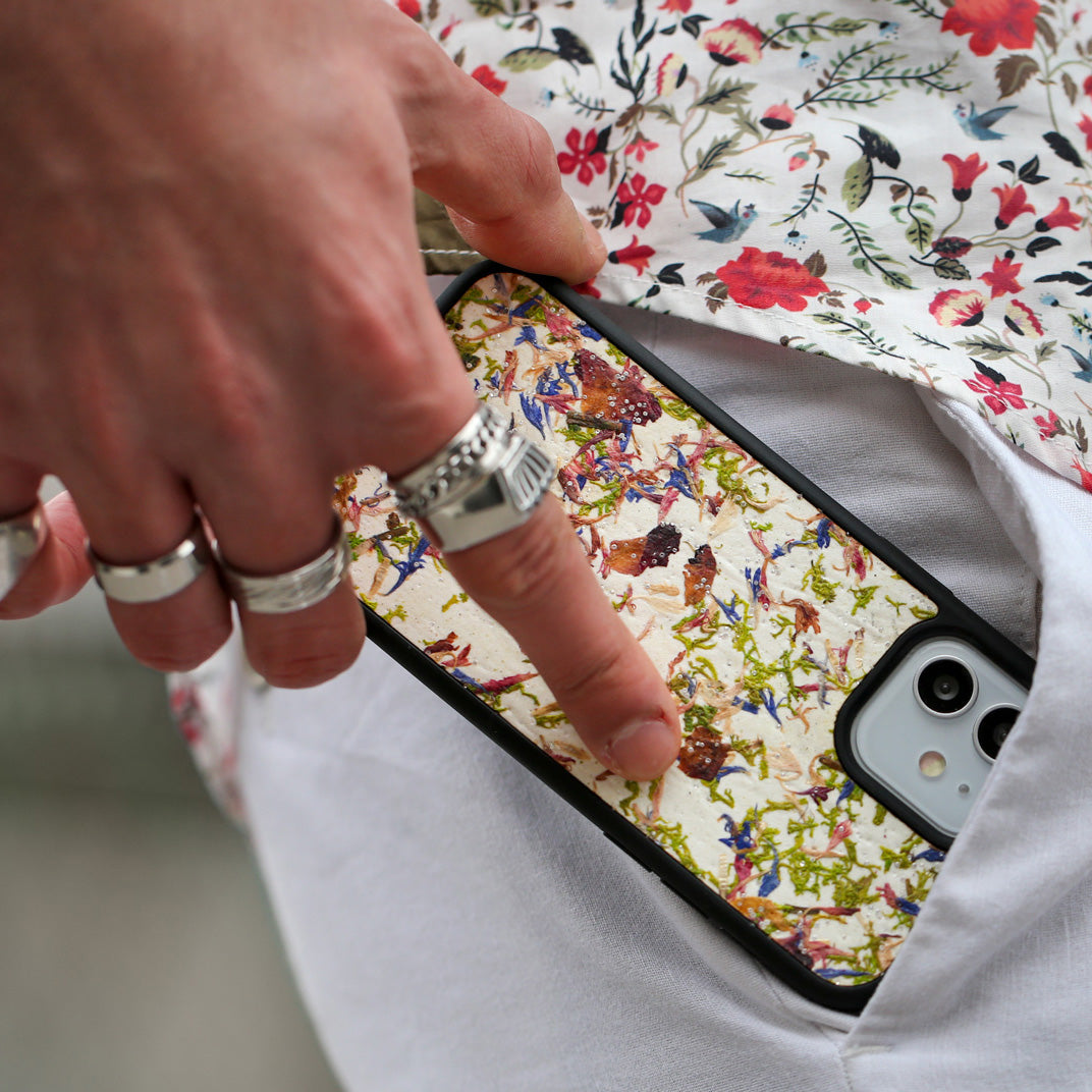 Un homme range son téléphone dans sa poche. Il a une belle main ornée de bague sur chacun de ses doigts. Le téléphone est vêtu d’une coque fleurie Hibisca exceptionnelle. La coque fleurie du téléphone a pour façade de multiples fleurs. Nous pouvons y apercevoir du blanc. Elle est aussi parsemée de multiple cristaux de chez Swarovski.  Cet homme porte également une très jolie chemise blanche avec des motifs fleuris rouges et verts.