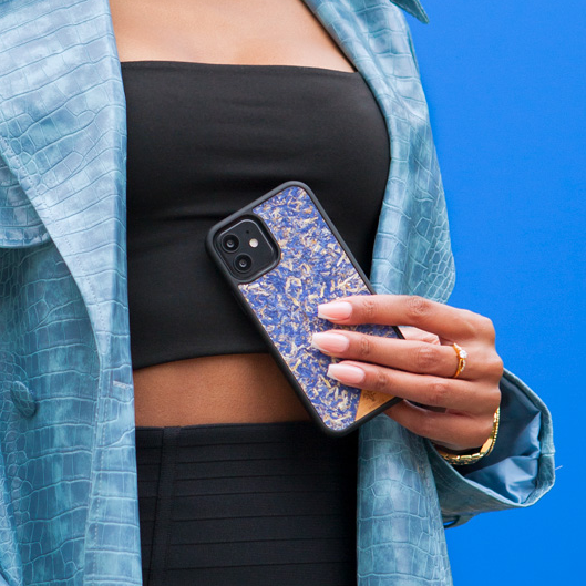 Une jolie femme capverdienne au regard charismatique tient son téléphone contre son torse. Son téléphone est vêtu d’une coque fleurie Hibisca. Elle a pour façade des fleurs de bleuets compressées. La coque est de couleur bleu. La femme a une belle main avec un vernis artistique sur ses ongles. Elle porte également un manteau bleu élégant avec un effet peau de crocodile avec une tenue moulante noire à l’intérieur.