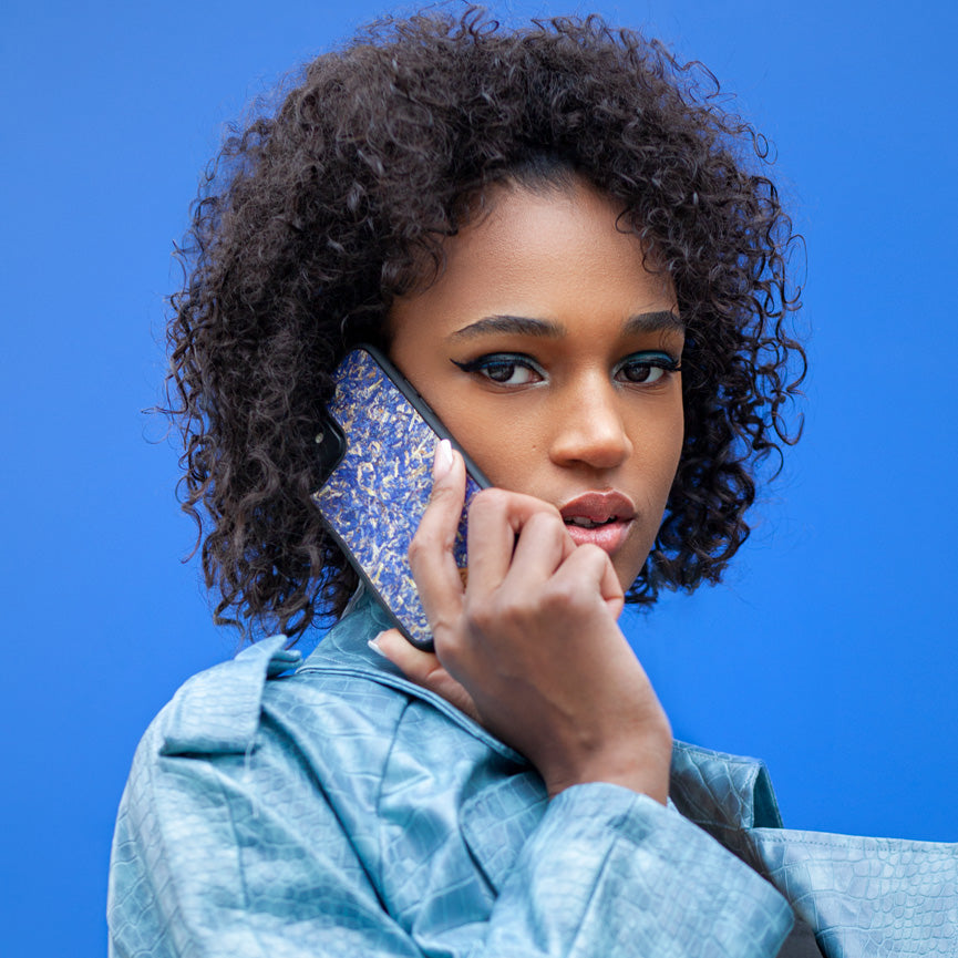 Une très jolie femme capverdienne au regard charismatique tient son téléphone contre son oreille. Son téléphone est vêtu d’une coque fleurie Hibisca. La coque est exceptionnelle. Elle a pour façade des fleurs de bleuets compressées très jolie à voir. La coque est donc de couleur bleu. La femme tenant le téléphone a une belle main avec un vernis artistique sur ses ongles. Elle porte également un manteau bleu élégant avec un effet peau de crocodile avec une tenue moulante noire à l’intérieur.