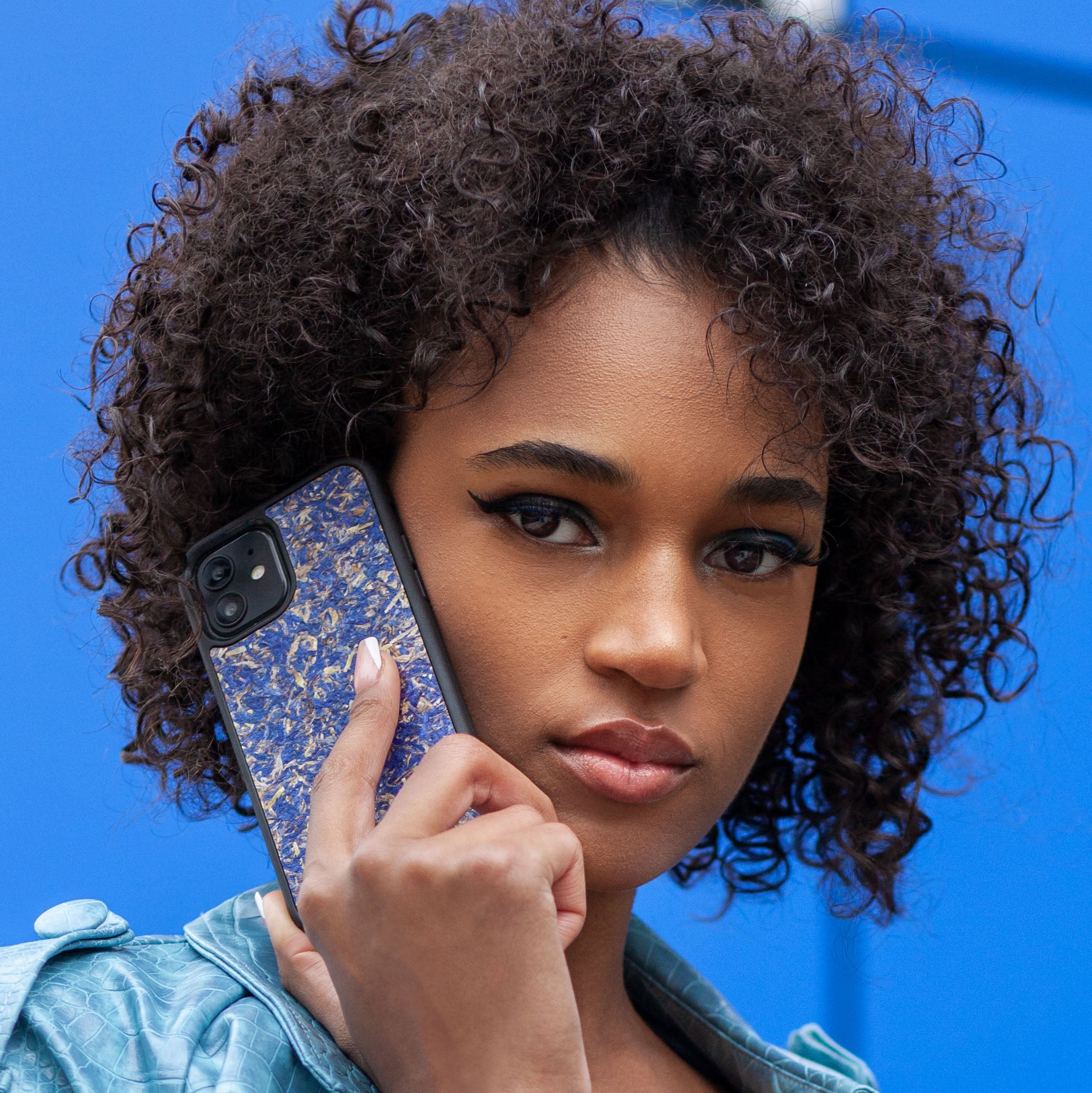 Une très jolie femme capverdienne au regard charismatique tient son téléphone contre son oreille. Son téléphone est vêtu d’une coque fleurie Hibisca. La coque est exceptionnelle. Elle a pour façade des fleurs de bleuets compressées très jolie à voir. La coque est donc de couleur bleu. La femme tenant le téléphone a une belle main avec un vernis artistique sur ses ongles. Elle porte également un manteau bleu élégant avec un effet peau de crocodile avec une tenue moulante noire à l’intérieur.