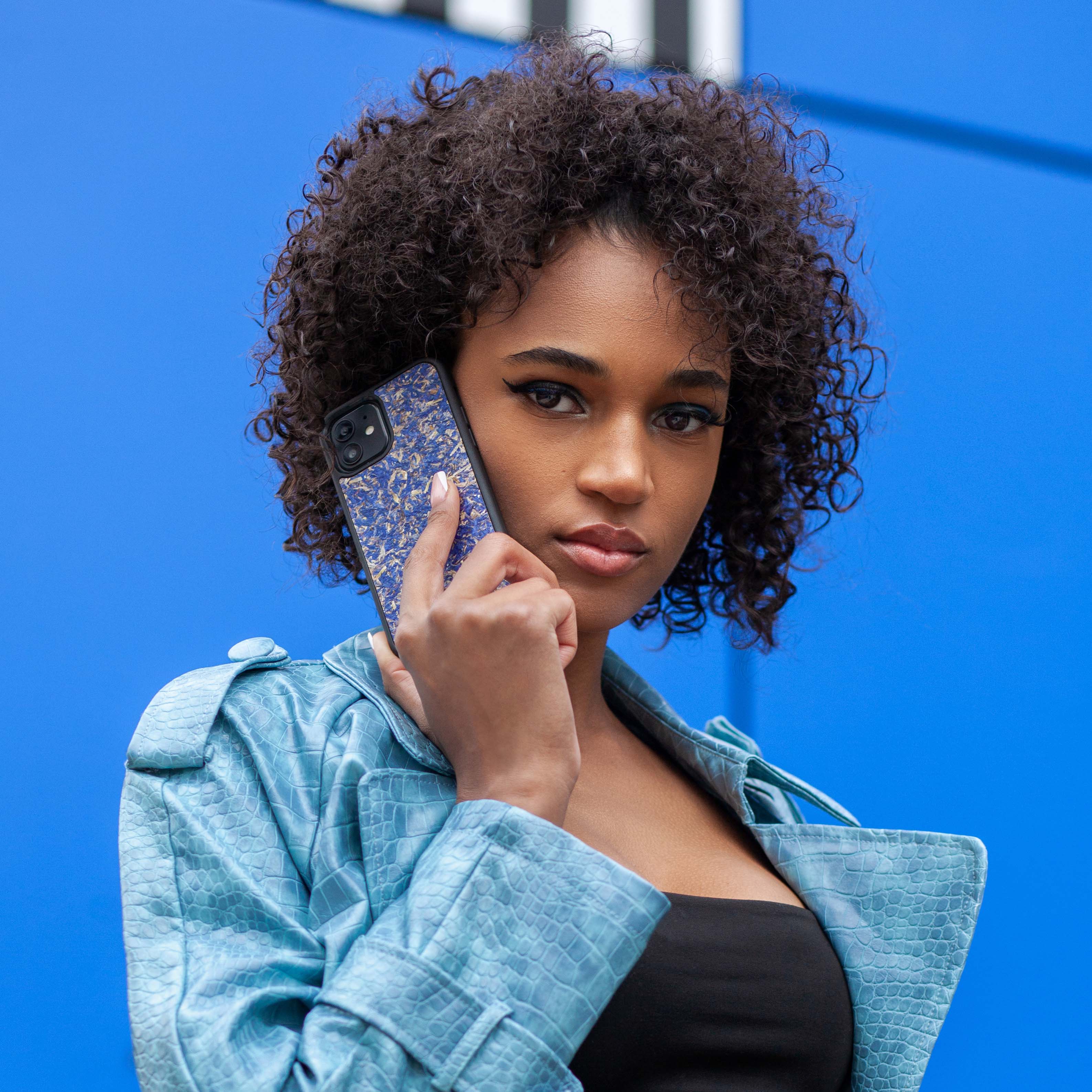 Une très jolie femme capverdienne au regard charismatique tient son téléphone contre son oreille. Son téléphone est vêtu d’une coque fleurie Hibisca. La coque est exceptionnelle. Elle a pour façade des fleurs de bleuets compressées très jolie à voir. La coque est donc de couleur bleu. La femme tenant le téléphone a une belle main avec un vernis artistique sur ses ongles. Elle porte également un manteau bleu élégant avec un effet peau de crocodile avec une tenue moulante noire à l’intérieur.