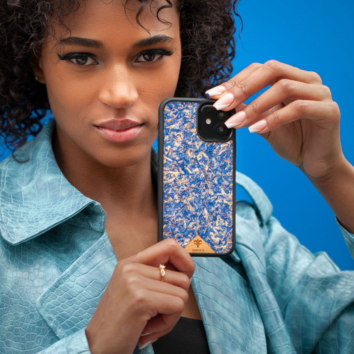 Une très jolie femme capverdienne au regard charismatique tient son téléphone contre son oreille. Son téléphone est vêtu d’une coque fleurie Hibisca. La coque est exceptionnelle. Elle a pour façade des fleurs de bleuets compressées très jolie à voir. La coque est donc de couleur bleu. La femme tenant le téléphone a une belle main avec un vernis artistique sur ses ongles. Elle porte également un manteau bleu élégant avec un effet peau de crocodile avec une tenue moulante noire à l’intérieur.