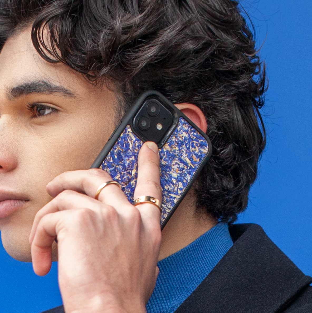 Un très bel homme au regard charismatique tient son téléphone contre son torse. Son téléphone est vêtu d’une coque Hibisca. La coque est exceptionnelle. Elle a pour façade des fleurs de bleuets compressées très jolie à voir. La coque est donc de couleur bleu. L’homme tenant le téléphone a une belle main ornée de bague sur chacun de ses doigts. Il porte également un très beau costume noir avec un tee-shirt élégant bleu rois.