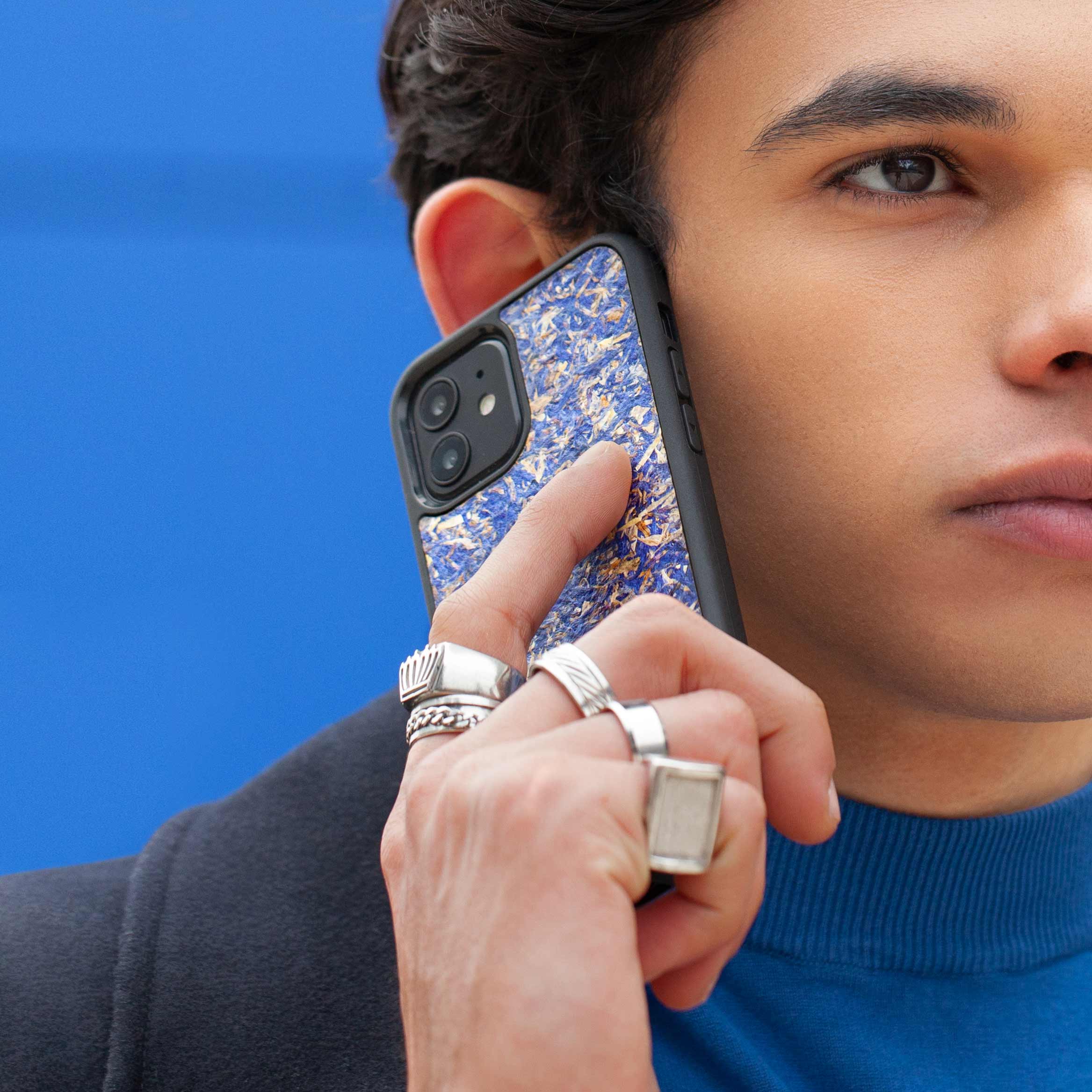 Un très bel homme au regard charismatique tient son téléphone contre son torse. Son téléphone est vêtu d’une coque Hibisca. La coque est exceptionnelle. Elle a pour façade des fleurs de bleuets compressées très jolie à voir. La coque est donc de couleur bleu. L’homme tenant le téléphone a une belle main ornée de bague sur chacun de ses doigts. Il porte également un très beau costume noir avec un tee-shirt élégant bleu rois.