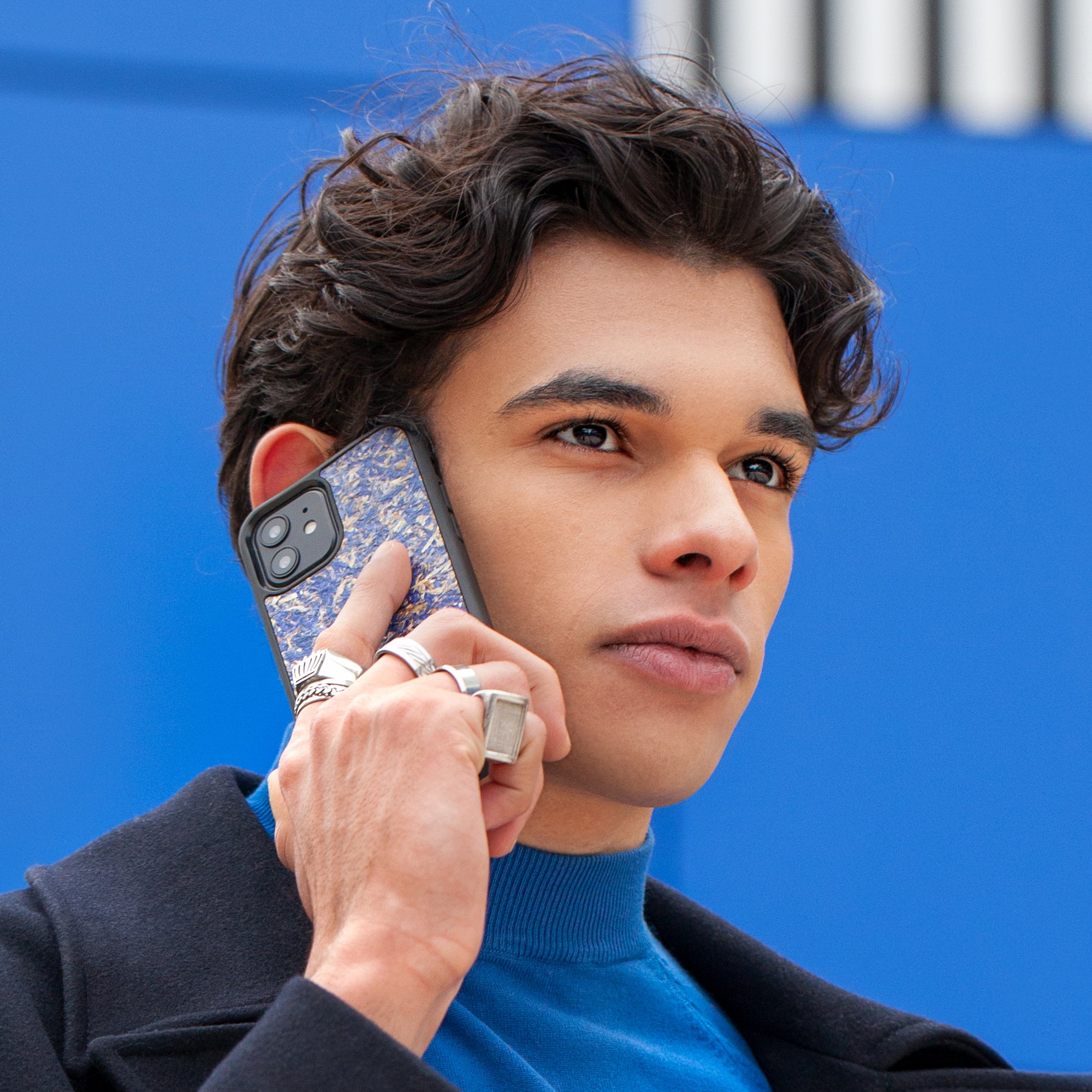 Un très bel homme au regard charismatique tient son téléphone contre son torse. Son téléphone est vêtu d’une coque Hibisca. La coque est exceptionnelle. Elle a pour façade des fleurs de bleuets compressées très jolie à voir. La coque est donc de couleur bleu. L’homme tenant le téléphone a une belle main ornée de bague sur chacun de ses doigts. Il porte également un très beau costume noir avec un tee-shirt élégant bleu rois.