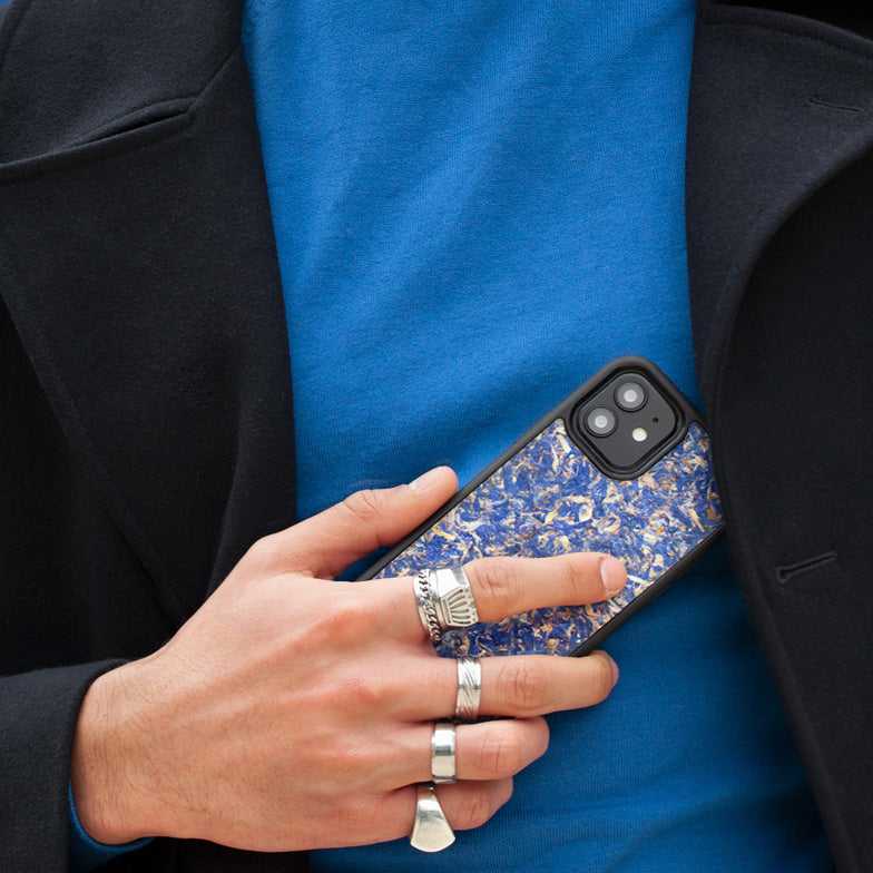 Un très bel homme au regard charismatique tient son téléphone contre son torse. Son téléphone est vêtu d’une coque fleurie Hibisca. La coque est exceptionnelle. Elle a pour façade des fleurs de bleuets compressées très jolie à voir. La coque est donc de couleur bleu. L’homme tenant le téléphone a une belle main ornée de bague sur chacun de ses doigts. Il porte également un très beau costume noir avec un tee-shirt élégant bleu rois.