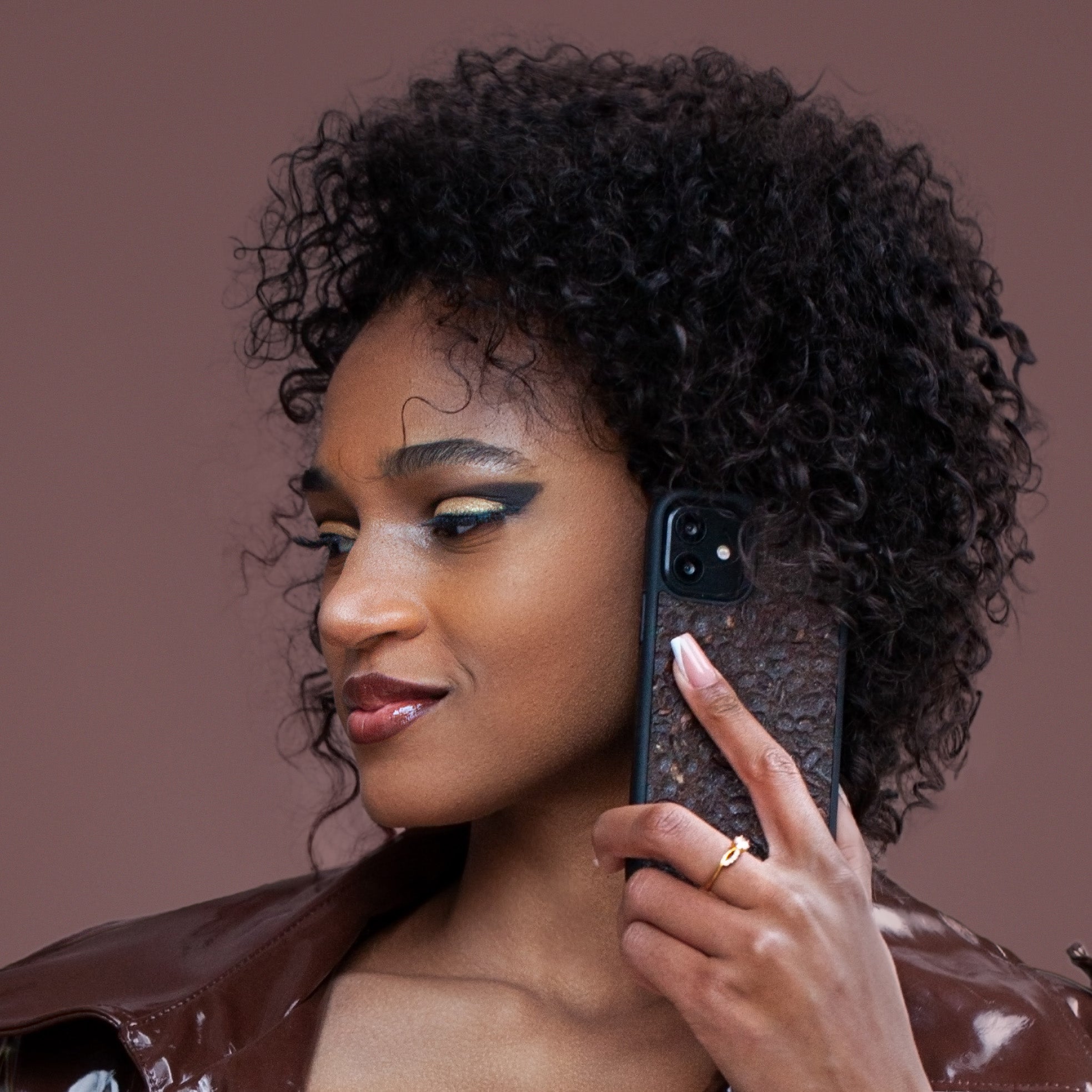 Une très jolie femme capverdienne au regard charismatique tient son téléphone contre son oreille. Son téléphone est vêtu d’une coque odorante et nervurée Hibisca. La coque odorante et nervurée est exceptionnelle. Elle a pour façade du café Arabica compressé d’une très belle façon. La coque odorante et nervurée est de couleur marron. La femme tenant le téléphone a une belle main avec un vernis
