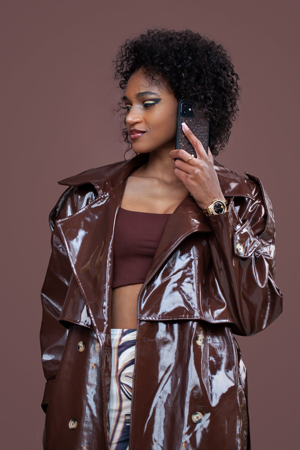 Une très jolie femme capverdienne au regard charismatique tiens son téléphone contre son oreille. Son téléphone est vêtu d’une coque Hibisca. La coque est exceptionnelle. Elle a pour façade du café Arabica compressé d’une très belle façon . La coque est de couleur marron. La femme tenant le téléphone a une belle main avec un vernis artistique sur ses ongles. Elle porte également une très belle tenue marron se mélangeant complètement au décors qui est lui aussi de couleur marron.