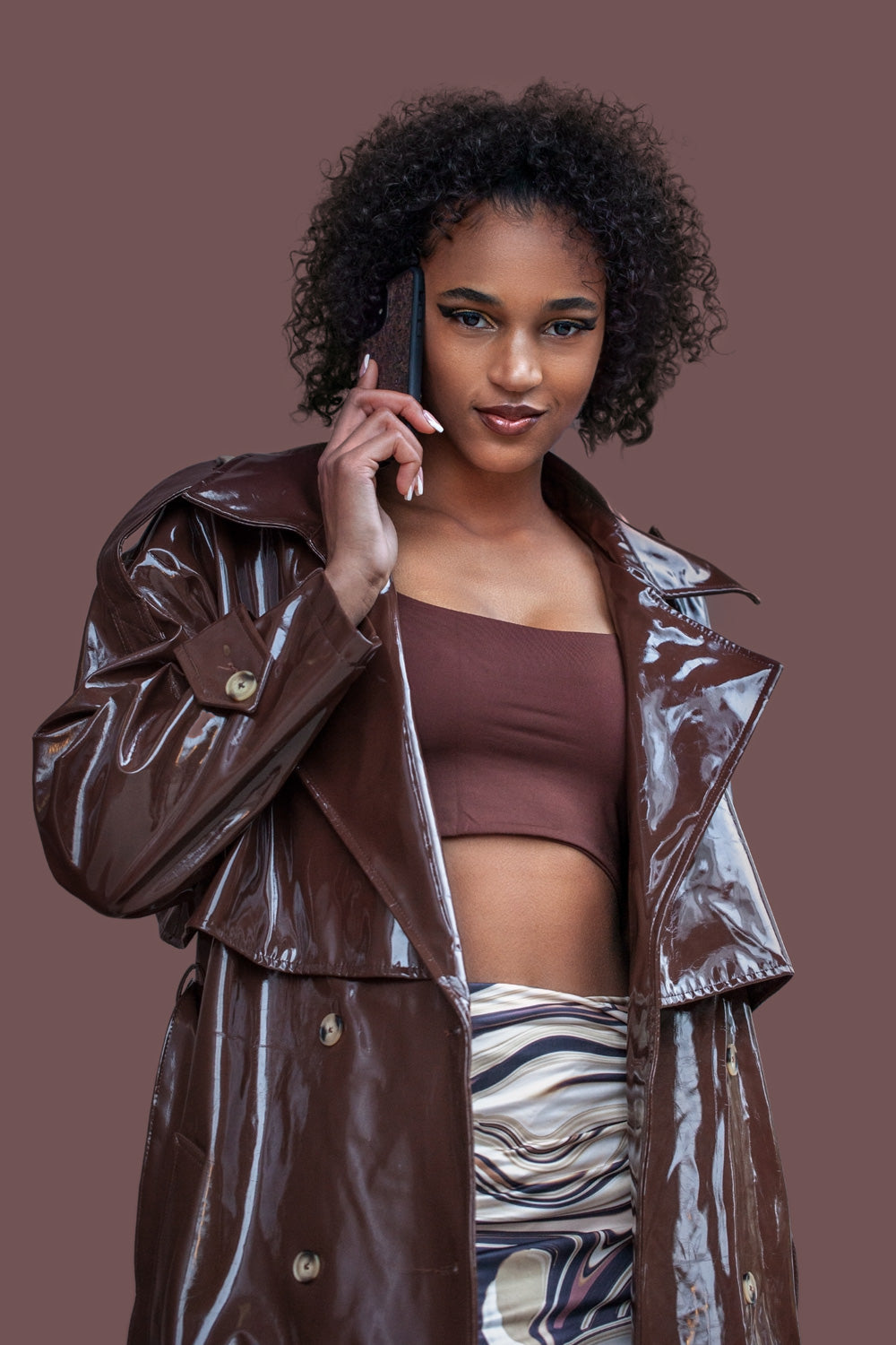 Une très jolie femme capverdienne au regard charismatique tiens son téléphone contre son oreille. Son téléphone est vêtu d’une coque Hibisca. La coque est exceptionnelle. Elle a pour façade du café Arabica compressé d’une très belle façon . La coque est de couleur marron. La femme tenant le téléphone a une belle main avec un vernis artistique sur ses ongles. Elle porte également une très belle tenue marron se mélangeant complètement au décors qui est lui aussi de couleur marron.