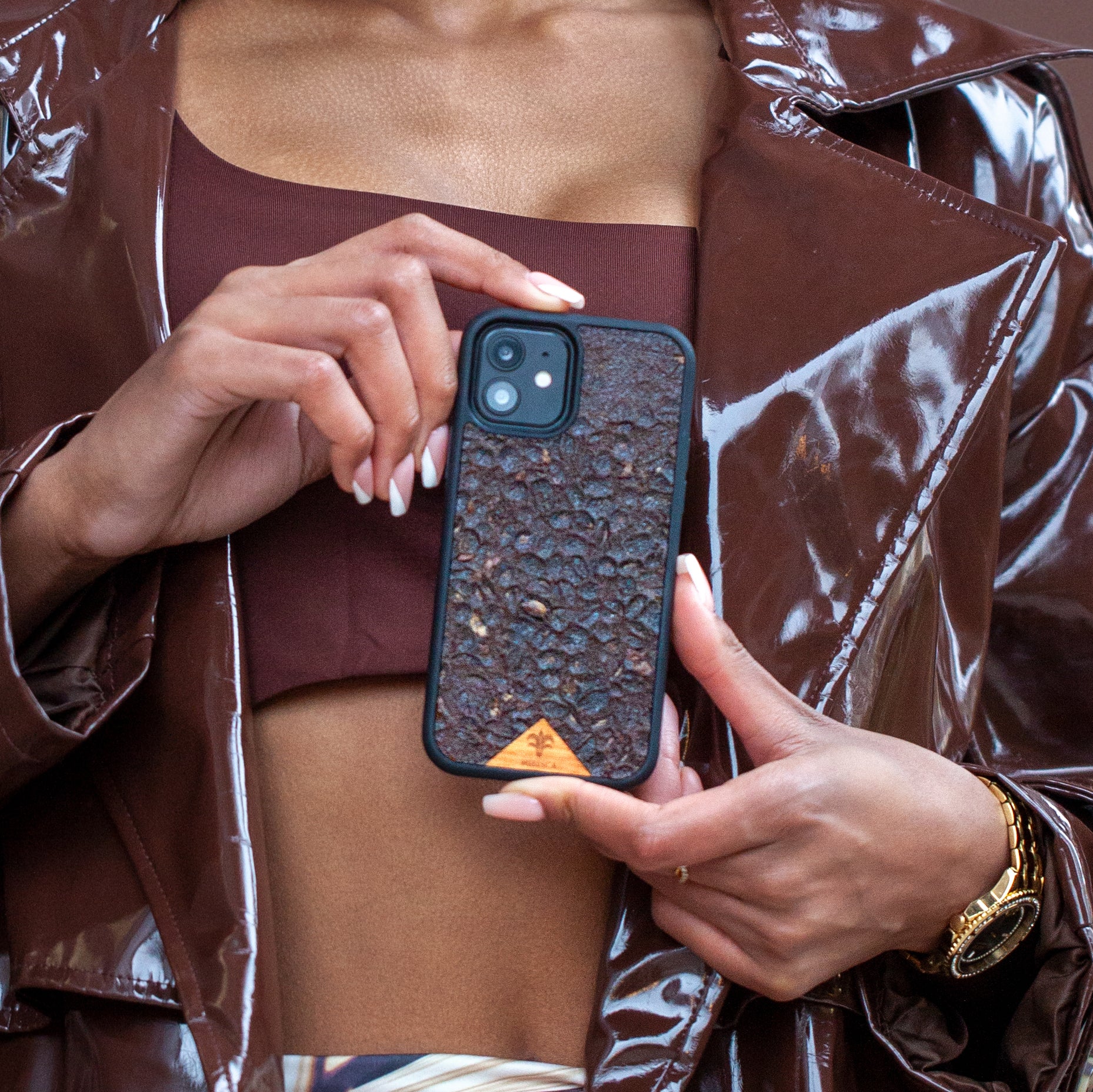 Une très jolie femme capverdienne tient son téléphone avec ses 2 mains d’une très belle façon. Son téléphone est vêtu d’une coque écoresponsable Hibisca odorante. La coque est nervurée. Elle a pour façade du café Arabica compressé d’une très belle façon. La coque est de couleur marron. La femme tenant le téléphone a de belles mains avec un vernis artistique sur ses ongles. Elle porte également une très belle tenue marron se mélangeant complètement au décor qui est lui aussi de couleur marron.