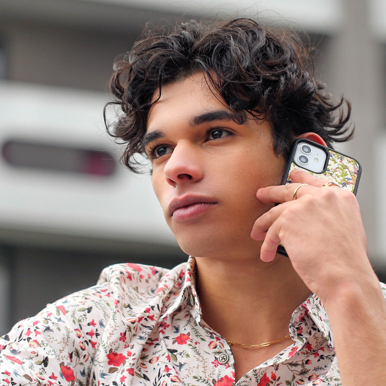 Un très bel homme au regard charismatique tient son téléphone contre son oreille. Son téléphone est vêtu d’une coque fleurie Hibisca. La coque fleurie est exceptionnelle. Elle a pour façade de multiples fleurs. Nous pouvons y apercevoir du blanc. Elle est aussi parsemée de multiple cristaux de chez Swarovski. L’homme tenant le téléphone a une belle main ornée de bague sur chacun de ses doigts. Il porte également une très jolie chemise blanche avec des motifs fleuris rouge et verts.