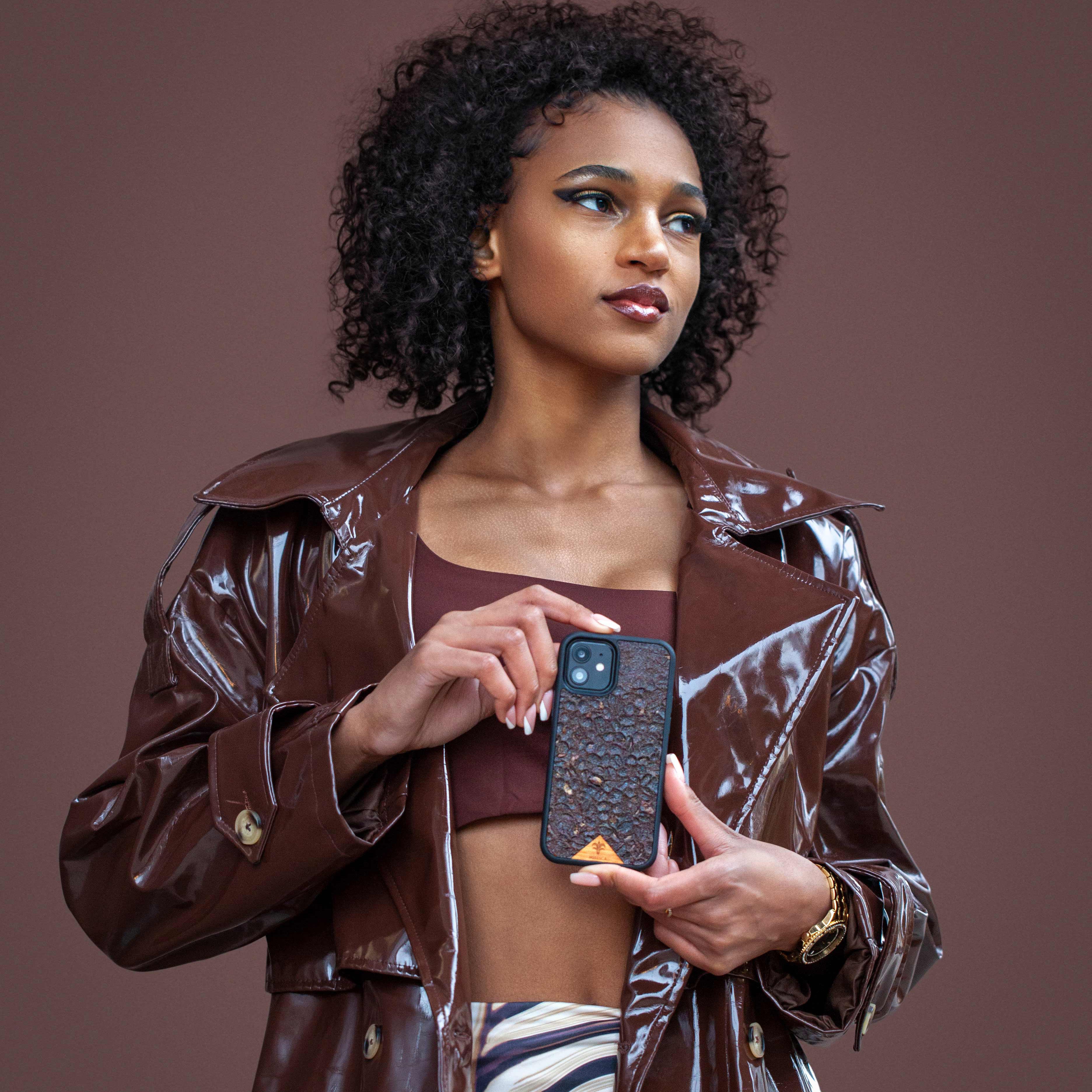 Une très jolie femme capverdienne tient son téléphone avec ses 2 mains d’une très belle façon. Son téléphone est vêtu d’une coque écoresponsable Hibisca odorante. La coque est nervurée. Elle a pour façade du café Arabica compressé d’une très belle façon. La coque est de couleur marron. La femme tenant le téléphone a de belles mains avec un vernis artistique sur ses ongles. Elle porte également une très belle tenue marron se mélangeant complètement au décor qui est lui aussi de couleur marron.  