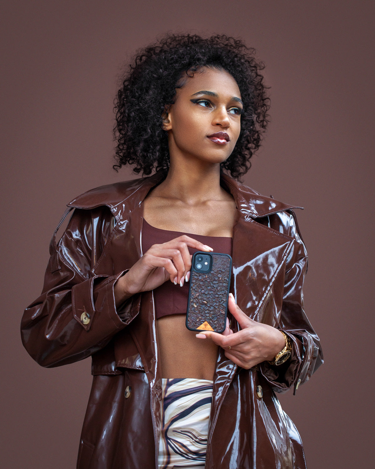 Une très jolie femme capverdienne tiens son téléphone avec ses 2 mains d’une très belle façon. Son téléphone est vêtu d’une coque Hibisca. La coque est exceptionnelle. Elle a pour façade du café Arabica compressé d’une très belle façon . La coque est de couleur marron. La femme tenant le téléphone a de belles mains avec un vernis artistique sur ses ongles. Elle porte également une très belle tenue marron se mélangeant complètement au décors qui est lui aussi de couleur marron.  